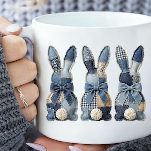 Può includere: Tazza in ceramica bianca con tre disegni di coniglietti patchwork in denim. Ogni coniglietto ha un fiocco blu e una coda a pompon beige. I coniglietti sono realizzati con vari motivi in denim.