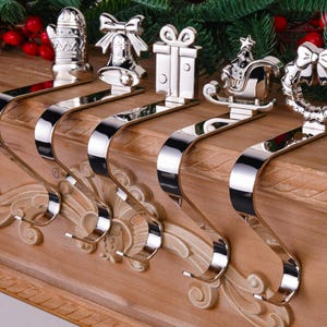 Può includere: Un set di cinque portacandele natalizi color argento. Ogni portacandele presenta un design festivo diverso: un guanto, una campana, una confezione regalo, una slitta con un albero e una ghirlanda. I portacandele sono esposti su un caminetto di legno.