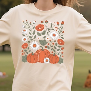 May include: A beige t-shirt featuring a fall-themed graphic. The design includes three orange pumpkins, white daisies, and red poppies, surrounded by green leaves and stems. The shirt has short sleeves and a crew neck.