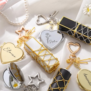 May include: An assortment of personalized keychains and compact mirrors in gold, silver, and black. The keychains feature heart, star, and rectangular shapes, some with names like "Elaine" and "Sophia". The compact mirrors have a decorative net-like design.