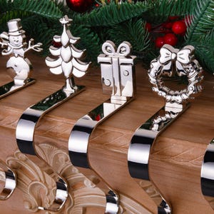 May include: A collection of silver Christmas stocking holders with festive designs, including a snowman, Christmas tree, gift, wreath, and snowflake. The holders are displayed on a wooden surface with Christmas greenery in the background.