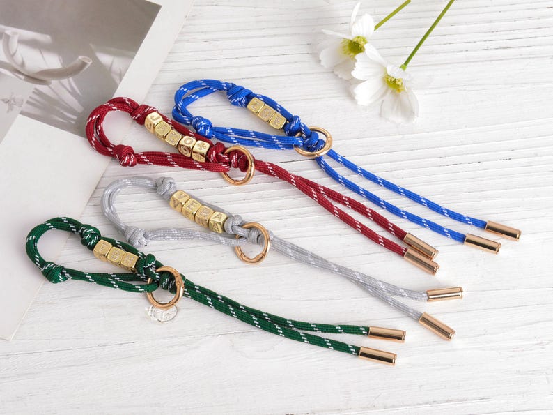 May include: Four keychains in red, blue, green, and gray with gold-colored hardware. Each keychain has a braided cord with a metallic end and a ring. Some keychains have lettered beads.