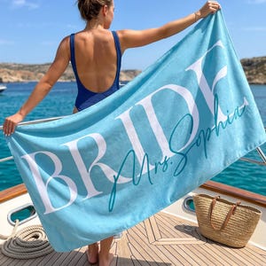 May include: A light blue rectangular beach towel featuring the word BRIDE in large white capital letters and the name Mrs. Sophia written in a teal cursive script font across the centre of the fabric surface.