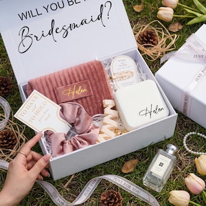 Könnte beinhalten: Eine weiße Geschenkbox mit der Aufschrift "Will you be Bridesmaid?" enthält ein rosa Handtuch, ein Scrunchie, Haarspangen und eine kleine weiße Box mit dem Namen "Helen". Eine Parfümflasche und eine Karte mit der Aufschrift "To have and to hold your hair back" sind ebenfalls enthalten.