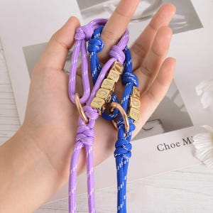 May include: Two dog collars, one lavender and one blue, are held in a hand. Each collar has a gold-colored ring and gold-colored lettered cubes spelling out a name. The collars are made of a woven rope material.