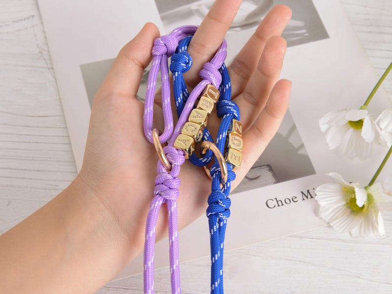 May include: Two dog collars, one lavender and one blue, are held in a hand. Each collar has a gold-colored ring and gold-colored lettered cubes spelling out a name. The collars are made of a woven rope material.