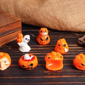 May include: A collection of Halloween-themed figurines, including orange pumpkins with carved faces, a white ghost holding a pumpkin, and a pumpkin with a skull inside. The figurines are arranged on a dark wooden surface.