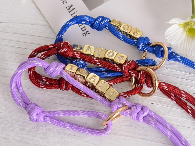 May include: Close-up of three dog collars in blue, red, and purple. Each collar features a rope design with gold-colored cube beads and a metal ring. The blue collar has white accents, the red has white accents, and the purple has a star pattern.