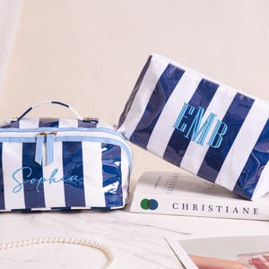 Könnte beinhalten: Zwei gestreifte Kosmetiktaschen in Blau und Weiß. Eine Tasche hat einen Griff und den Namen "Sophia" in Schreibschrift. Die andere Tasche hat die Initialen "EMB" aufgestickt. Beide Taschen stehen auf einer weißen Oberfläche.