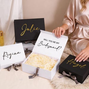 Puede incluir: Cuatro cajas de regalo, dos negras y dos blancas, con texto dorado o negro. La caja blanca está abierta, llena de papel triturado, y dice "Will you be my Bridesmaid?" Las otras cajas tienen los nombres: "Julie", "Regina" y "Hazel".