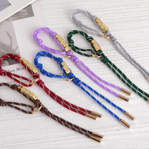 May include: A collection of colorful keychains with braided cords in brown, burgundy, blue, purple, green, and gray. Each keychain features a gold-colored metal accent with a loop and a set of small, square beads. The ends are finished with gold-colored metal caps.