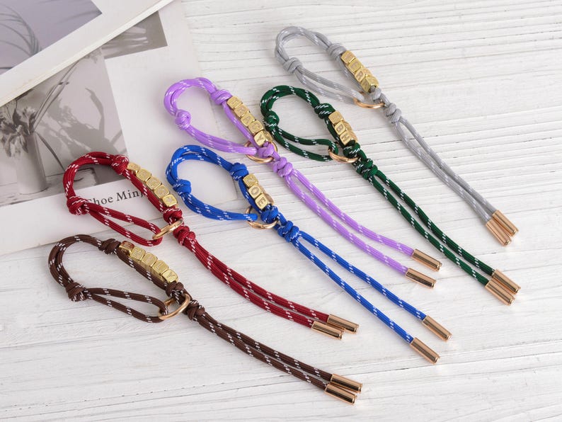 May include: A collection of colorful keychains with braided cords in brown, burgundy, blue, purple, green, and gray. Each keychain features a gold-colored metal accent with a loop and a set of small, square beads. The ends are finished with gold-colored metal caps.