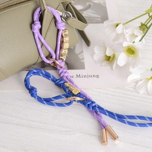 May include: A close-up of a purse strap in shades of purple and blue, with gold-colored accents. The strap is made of braided rope and features small gold letter beads spelling a name. White flowers and a purse are in the background.