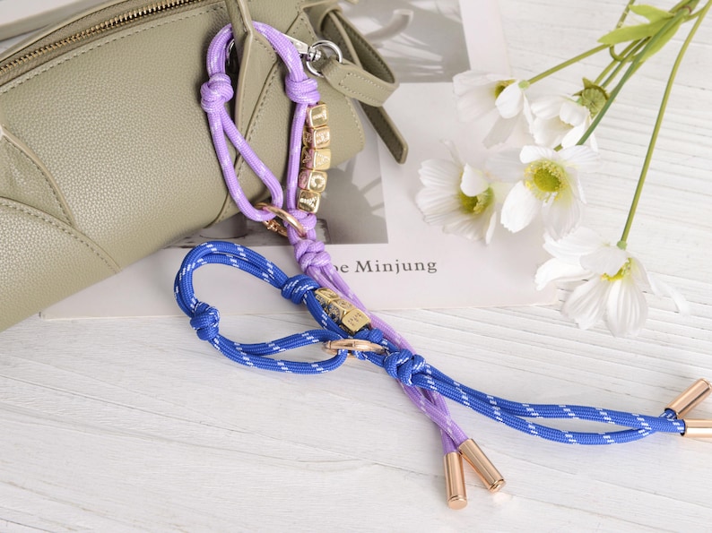 May include: A close-up of a purse strap in shades of purple and blue, with gold-colored accents. The strap is made of braided rope and features small gold letter beads spelling a name. White flowers and a purse are in the background.