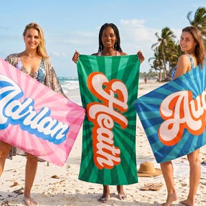 May include: Three rectangular beach towels featuring a sunburst pattern. The left towel is pink with the white text Adrian. The center towel is green with the orange text Heath. The right towel is blue with the orange text August.