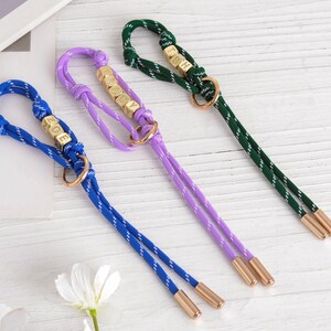 May include: Three colorful dog leashes in blue, purple, and green. Each leash features a rope design with gold-colored accents, including small lettered cubes spelling out a word. The leashes have a loop at the top and two dangling cords.