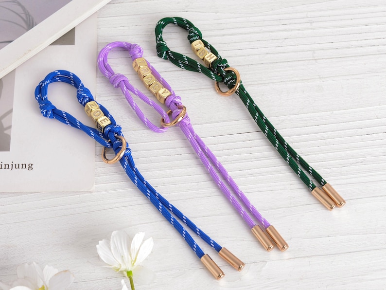 May include: Three colorful dog leashes in blue, purple, and green. Each leash features a rope design with gold-colored accents, including small lettered cubes spelling out a word. The leashes have a loop at the top and two dangling cords.