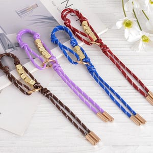 May include: Four keychains in brown, purple, blue, and red. Each keychain has a braided cord, gold-colored metal accents, and a small ring. The keychains are arranged on a white surface.