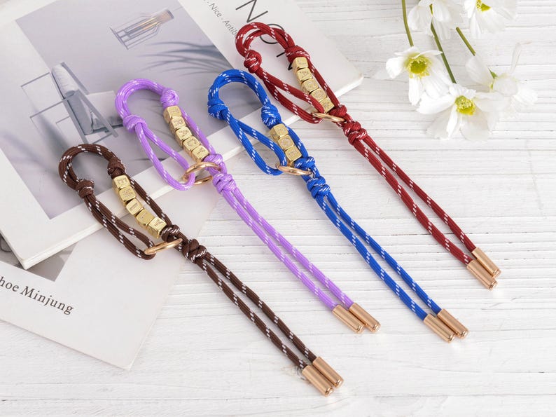 May include: Four keychains in brown, purple, blue, and red. Each keychain has a braided cord, gold-colored metal accents, and a small ring. The keychains are arranged on a white surface.