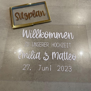 Könnte beinhalten: Ein klares Hochzeitszeichen aus Acryl mit weißer Schrift. Auf dem Schild steht "Willkommen zu unserer Hochzeit Emilia & Matteo 27. Juni 2023." Ein goldgerahmtes Glasschild mit dem Wort "Sitzplan" befindet sich darüber.