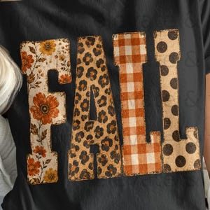 Könnte beinhalten: Ein schwarzes Kurzarm-T-Shirt mit dem Wort FALL in großen Blockbuchstaben. Jeder Buchstabe hat ein eigenes Muster: das F zeigt orangefarbene Blumen, das A ein Leopardenmuster, das L ein orangefarbenes Vichy-Karo und das L braune Punkte.