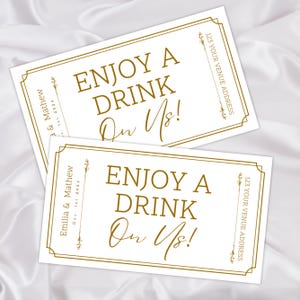 Op de afbeelding: Twee witte rechthoekige kaarten met gouden tekst die luidt "ENJOY A DRINK On Us!". De kaarten hebben gouden randen en bevatten de namen "Emilia & Mathew" en de datum "Dec. 1st 2024". De kaarten bevatten ook de tekst "123 YOUR VENUE ADDRESS".