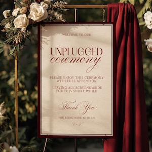 Op de afbeelding: Een ingelijst bord met bordeauxrode tekst met de tekst "WELCOME TO OUR UNPLUGGED ceremony". Het bord zegt ook "PLEASE ENJOY THIS CEREMONY WITH FULL ATTENTION" en "Thank You". Het bord wordt weergegeven op een gouden standaard met een bordeauxrode drape.