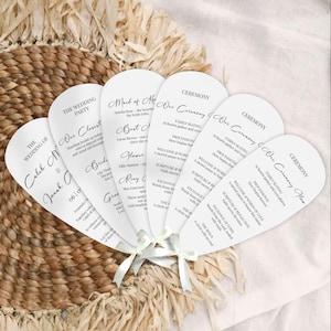 May include: A collection of white, fan-shaped wedding programs tied with ivory ribbons. Each program features elegant black script with details like the ceremony order, names, and dates. The programs rest on a woven, textured surface.