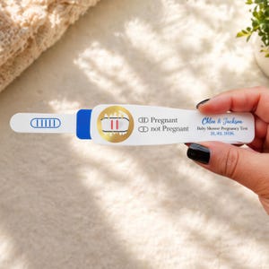 Könnte beinhalten: Ein weißer Schwangerschaftstest mit blauen und goldenen Akzenten. Der Test zeigt zwei rote Linien, was ein positives Ergebnis anzeigt. Der Text auf dem Test lautet "Pregnant" und "Chloe & Jackson Baby Shower Pregnancy Test 31, 03, 2026."