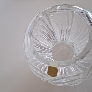 May include: A clear, round crystal vase with a spiral design. The vase has a wide opening and a thick base. The crystal reflects light, creating a sparkling effect. A small, gold-colored sticker is visible on the side.