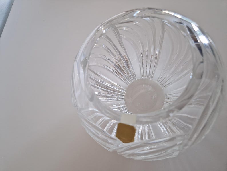 May include: A clear, round crystal vase with a spiral design. The vase has a wide opening and a thick base. The crystal reflects light, creating a sparkling effect. A small, gold-colored sticker is visible on the side.