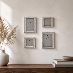 Può includere: Quattro cornici per foto in legno grigio di varie dimensioni sono montate su una parete di colore chiaro. Ogni cornice ha un interno grigio chiaro e una piccola puntina di colore scuro. Le cornici sono disposte in un motivo rettangolare. Un vaso con piante secche è a sinistra.