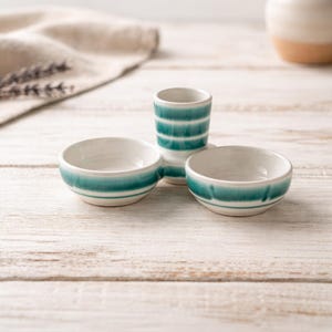 Può includere: Un set di tre piccole ciotole in ceramica e un bicchierino. Le ciotole e il bicchierino sono bianchi con strisce turchesi. Gli oggetti sono disposti su una superficie di legno chiaro.
