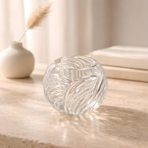 May include: A clear, round glass vase with a leaf-like pattern. The vase is sitting on a light-colored surface. A small white vase with a dried plant is in the background.