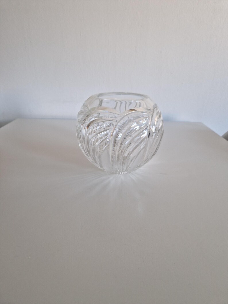 May include: A clear, round glass vase with a textured, leaf-like pattern. The vase is sitting on a white surface. The vase is likely used for decorative purposes.