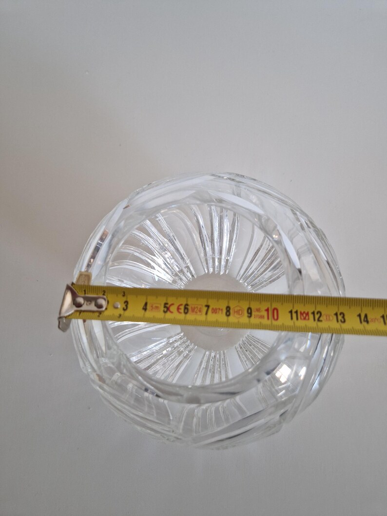 May include: A clear, round crystal bowl with a textured, swirling design. A yellow tape measure is placed across the bowl, showing a diameter of approximately 4.5 inches. The bowl is on a white background.
