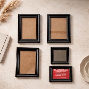 May include: A set of five black picture frames with a brown backing. The frames are arranged on a light-colored surface. One frame has a red label with white text. The frames vary in size, with the largest being rectangular and the smallest square.