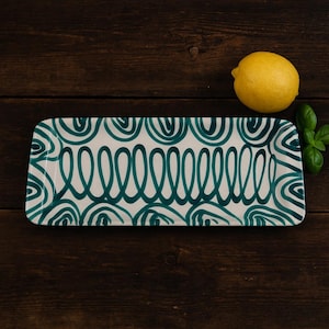Könnte beinhalten: Rechteckige Servierplatte mit weißem Hintergrund und tealfarbenem, abstraktem Design. Eine gelbe Zitrone und ein Basilikumzweig liegen auf einer dunklen Holzoberfläche neben der Platte. Das Muster der Platte zeigt eine Reihe verbundener Schlaufen und Halbkreise.