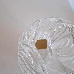 May include: Clear crystal bowl with a swirling, textured design. The bowl has a unique shape and is partially visible, showcasing the intricate cuts and patterns. A small, gold-colored label is attached to the bowl.