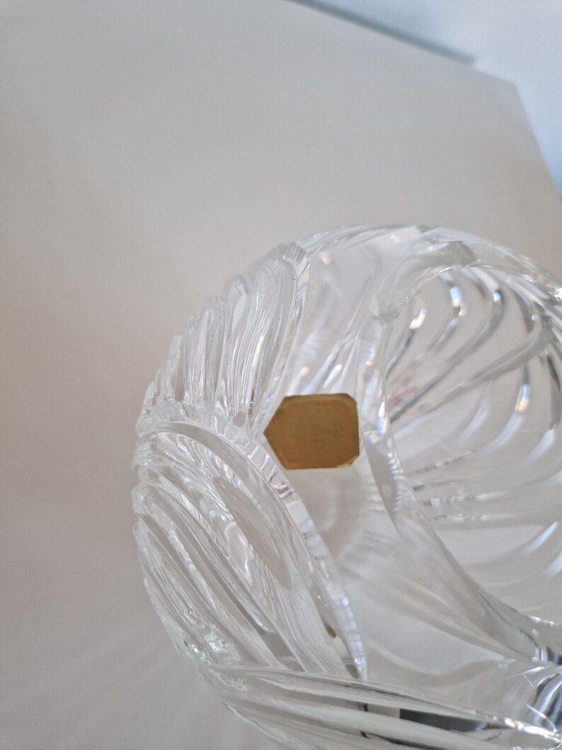 May include: Clear crystal bowl with a swirling, textured design. The bowl has a unique shape and is partially visible, showcasing the intricate cuts and patterns. A small, gold-colored label is attached to the bowl.