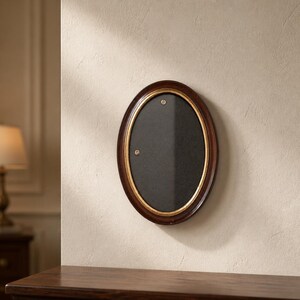 Peut inclure: Cadre photo ovale avec un cadre en bois marron foncé et un liseré intérieur doré. Le cadre a un intérieur noir et est monté sur un mur beige clair. Le cadre est vide et prêt pour une photo.