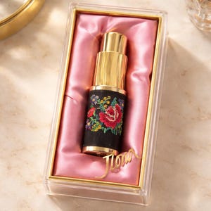 Può includere: Un flacone di profumo dorato con un motivo floreale ricamato nero, presentato in una scatola trasparente con fodera in raso rosa. Il ricamo presenta rose rosse e dettagli colorati. La parola "Flora" dorata è nella parte inferiore della scatola.