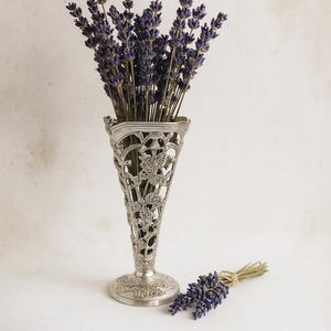 Könnte beinhalten: Eine silberfarbene, verzierte Vase gefüllt mit getrocknetem Lavendel. Die Vase hat ein detailliertes, durchbrochenes Design. Ein kleines Lavendelbüschel liegt neben der Vase. Der Lavendel hat einen violetten Farbton, der Hintergrund ist neutral.