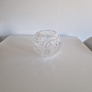 May include: A clear, spherical crystal vase with a textured, carved design. The vase sits on a white surface, reflecting light and creating shadows. The cut crystal pattern adds a decorative element to the piece.
