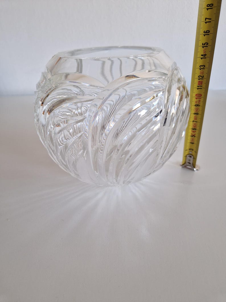 May include: A clear, round glass vase with a textured, swirling design. The vase is approximately 5 inches in diameter. The vase is sitting on a white surface.