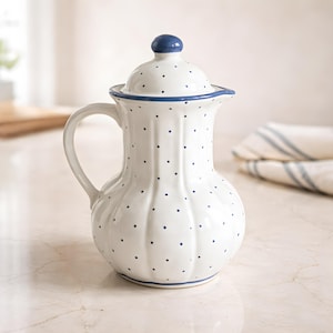 Puede incluir: Jarra de cerámica blanca con tapa, adornada con pequeños lunares azules. La jarra tiene un asa curva y un borde azul. La tapa está rematada con un pequeño pomo azul. Un artículo de cocina clásico.