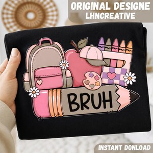 May include: A black t-shirt featuring a graphic print of a pink backpack, a red apple with a pink baseball cap, a box of crayons, a paint palette, and a large pencil with the text BRUH printed in black letters across the center of the pencil.