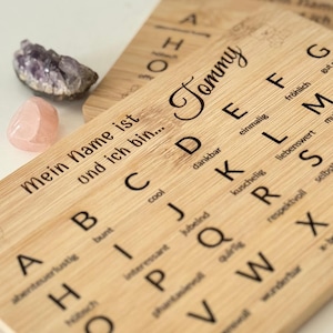 Könnte beinhalten: Zwei Holztafeln mit dem Alphabet und dem eingravierten Namen "Tommy". Die Tafeln zeigen das Alphabet mit deutschen Wörtern. Neben den Tafeln liegen ein kleiner Amethyst und ein rosa Kristall.