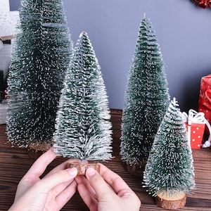 Peut inclure: Plusieurs petits sapins de Noël verts avec des pointes givrées blanches et des bases en bois. Les arbres de différentes tailles sont disposés sur une surface en bois. Les mains d'une personne tiennent l'un des arbres. Autres décorations de vacances.