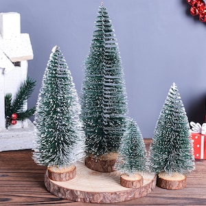 May include: A collection of miniature green Christmas trees with snow-like accents, displayed on wooden bases. The trees vary in size and are arranged on a wooden surface, with a decorative house and wrapped gifts in the background.